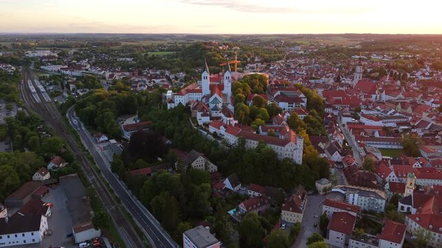 Drone footage of Freising, Bavaria, Germany, shows the Domberg with Freising Cathedral and St. George Church overlooking the old town skyline during a vivid, colorful sunset.