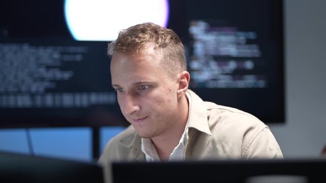A software developer focusing on programming desktop computer monitors in a corporate office workspace while a colleague works in the background.