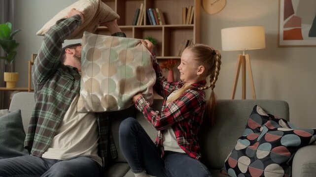 Playful Caucasian family dad man playing pillow fight little girl daughter home male parent happy kid funny battle at sofa guy father child schoolgirl fighting laughing together fun carefree weekend
