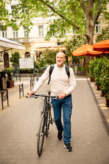 Fototapeta premium Happy man walking with a bicycle on a sunny city street, carrying a backpack outdoors