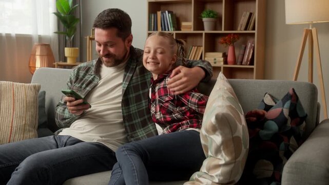 Caucasian male use mobile phone happy girl daughter jump at couch hugging daddy parent father man scrolling online kid family at home adoption smiling dad guy cuddling little child schoolgirl bonding