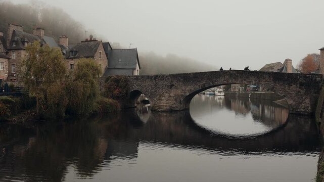 Historic stone arch bridge over the river in Dinan, France. Reflections of charming riverside houses and the bridge shimmer on calm water as pedestrians cross through the misty morning. A peaceful gli