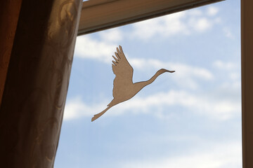 Fototapeta premium Paper cutout of a flying crane positioned against a window with blue sky and clouds visible, creating a serene indoor atmosphere