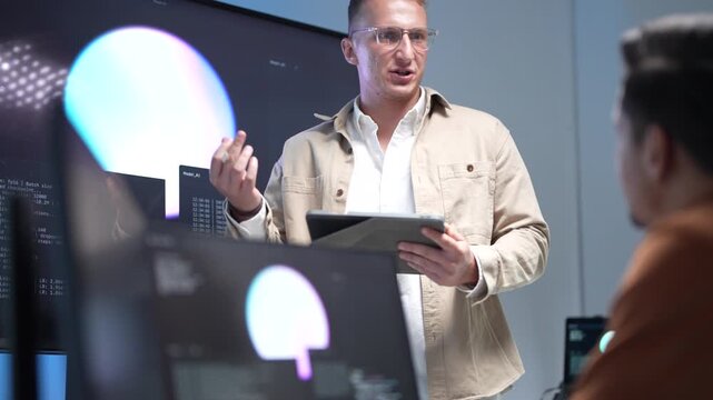 A senior IT specialist wearing glasses and holding a tablet while presenting machine learning workflows and artificial intelligence algorithms on a large screen.