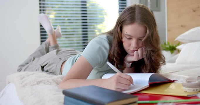 Teen girl lying on bed writing notes in open notebook with pen, studying with books