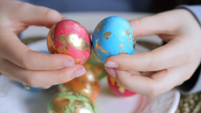 Hands hold decorated Easter eggs with gold foil. Colorful eggs sit on a white plate. Focus on hands and eggs in foreground. Background slightly blurred