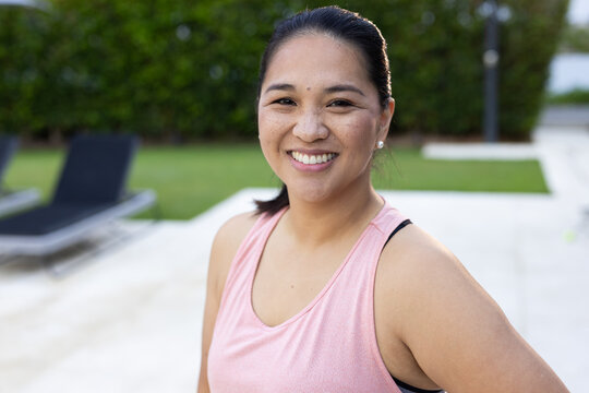 Mid-adult Asian woman smiling, posing on paved patio wearing pink tank top, showing black strap