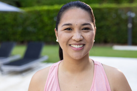 Asian female standing poolside on patio smiling at camera wearing pink sports top and pearl studs