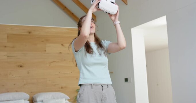 Woman 20s lifting white VR headset to eyes, adjusting fit by headboard in bedroom trying VR