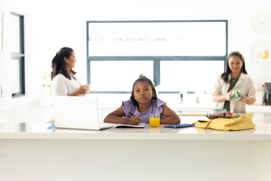 Diverse female family standing, child girl sitting and working on laptop at kitchen island
