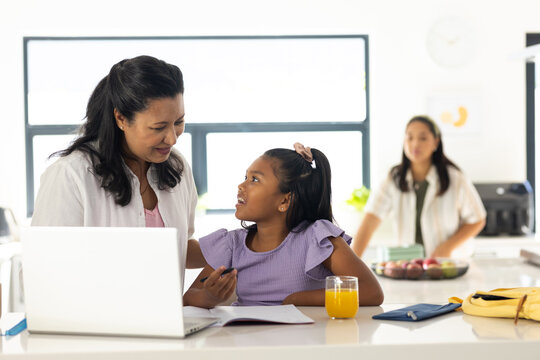 Diverse family leaning and helping youth girl studying at kitchen island with laptop, juice