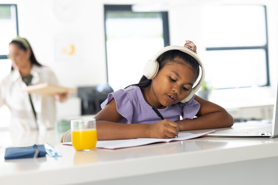 African American mom passing by as preteen daughter writing in notebook at home with white headset