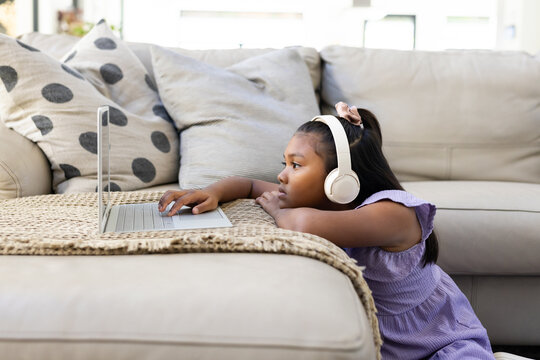 African American child lying by sofa in purple dress using laptop wearing headset, copy space