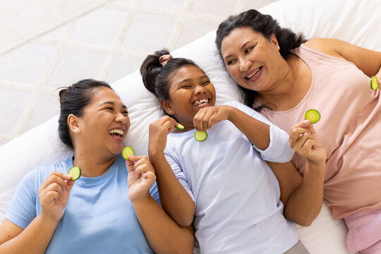 Three-generation female family lying on white cushion at home in pastel tops holding cucumbers