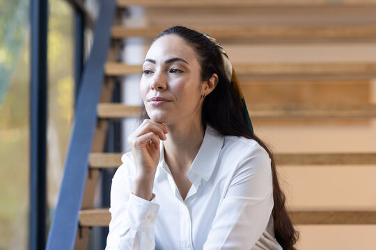 Woman sitting on wooden stairs by window wearing white blouse and patterned scarf gazing left