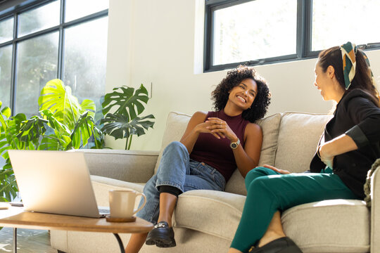 Female friends sitting and chatting on light-colored sofa in bright living room with open laptop