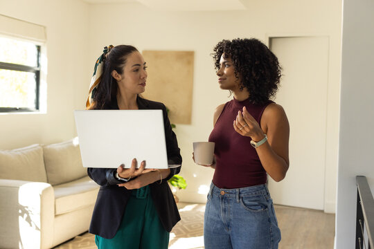 Two adult female colleagues standing and talking in bright living room holding laptop ceramic mug