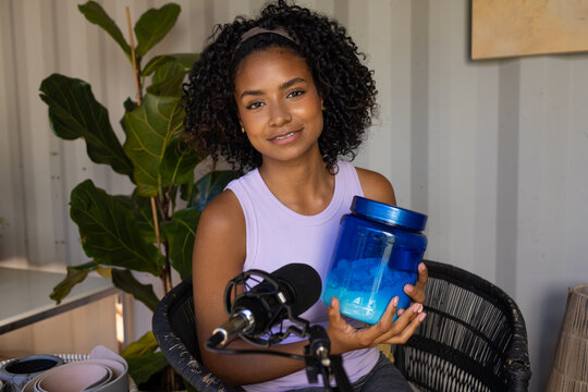 Female creator holding blue jar and speaking into microphone in home studio wearing lilac top