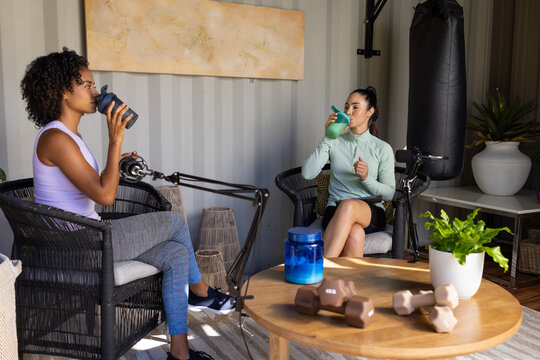Diverse female friends wearing sportswear chatting, sipping shakers in podcast nook with mic boom
