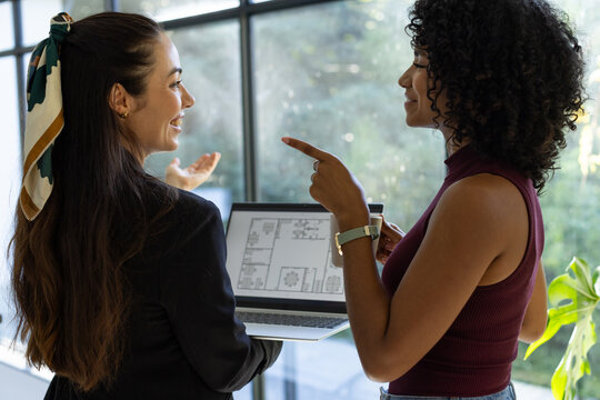Diverse female colleagues wearing patterned scarf discussing laptop floor plan in office by windows