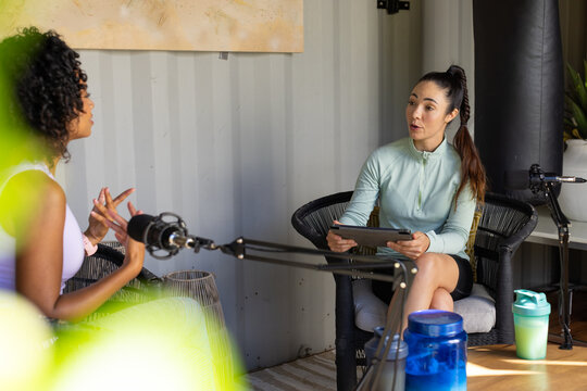 Diverse female interviewer and guest sitting in home studio wearing sport top using tablet and mics