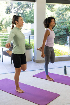 Diverse female friends in activewear doing quad stretch on purple mats on patio with water bottle