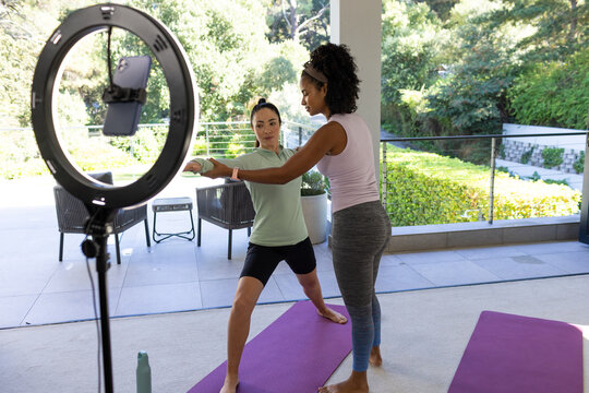 Diverse female instructor guiding student practicing yoga in sportswear on patio with ring light