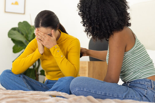 Female friends sitting on bed in bedroom, left covering face wearing mustard top, other comforting