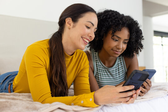 Diverse female friends lying on bed in bedroom, holding smartphones on plush blanket