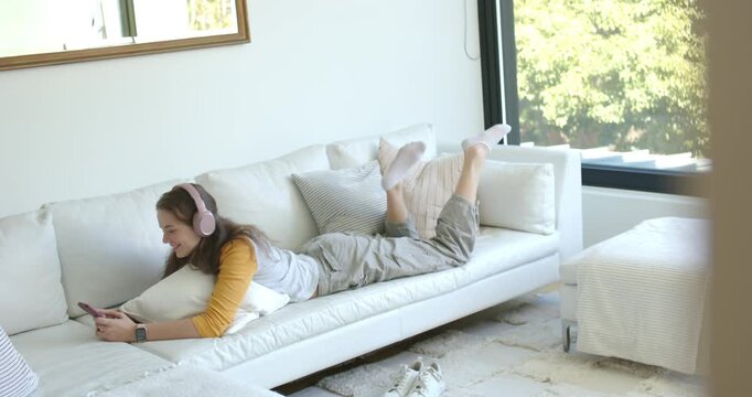 Woman lying on sofa listening on pink headphones, scrolling phone and kicking feet in living room