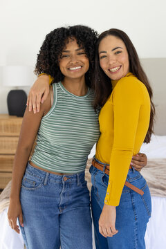 Diverse female friends standing close in bedroom, smiling at camera with bed, jeans and brown belt