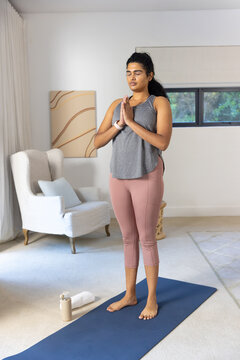 Woman practicing prayer pose in living room on blue mat wearing top, leggings with water bottle