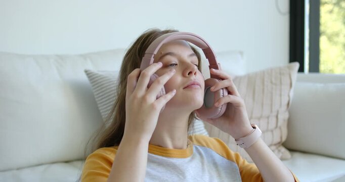 Woman leaning back on sofa grabbing pink headphones, placing them over ears and adjusting to listen