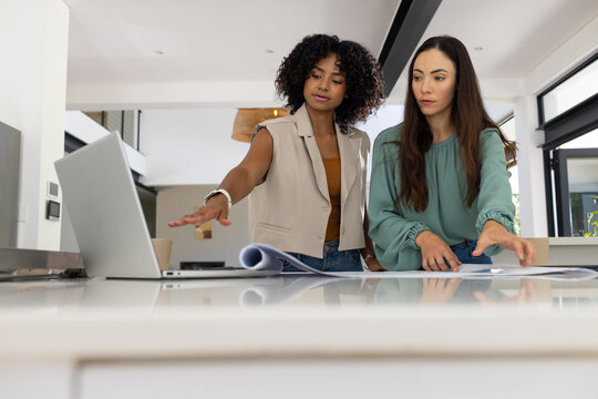 Diverse female colleagues standing at kitchen island reviewing blueprints and using open laptop