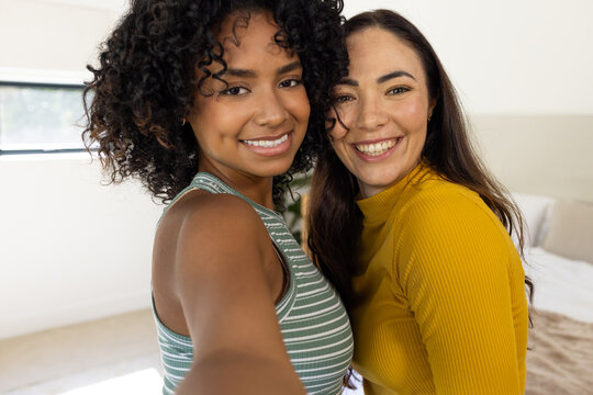 Female friends posing for selfie with smartphone in bedroom wearing striped tank and mustard top