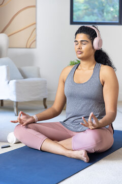 Indian adult woman meditating cross-legged on blue mat at home wearing gray tank and headphones