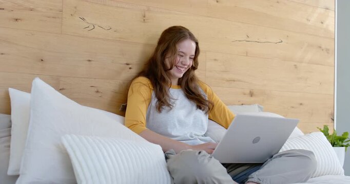Woman sitting cross-legged on bed scrolling and typing on laptop, reacting to funny message