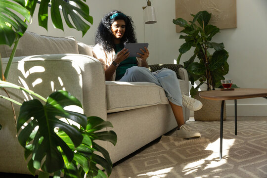 Indian woman sitting on sofa in sunny living room using tablet, sunlight casting large-leaf shadows