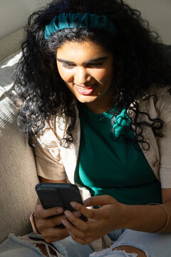 Woman sitting on light-colored couch in sunlight holding black smartphone and wearing teal headband