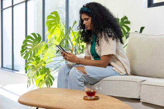 Indian woman sitting on sofa, scrolling smartphone by windows with Monstera, copy space