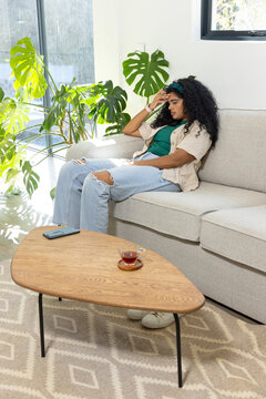 Woman reclining on sofa in living room holding forehead beside teacup and smartphone, copy space