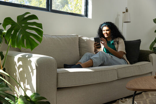 Adult woman sitting cross-legged right of beige sofa in living room, holding smartphone, copy space