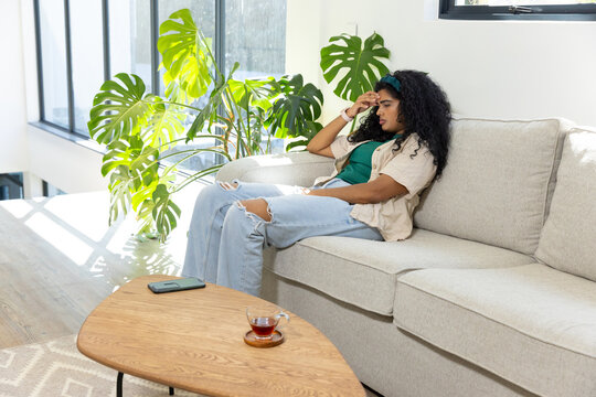 Indian woman sitting on right-sofa, holding head, in jeans, phone and tea on table, copy space