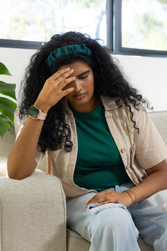 Indian woman sitting on sofa at home, holding head, wearing green headband and pink smartwatch