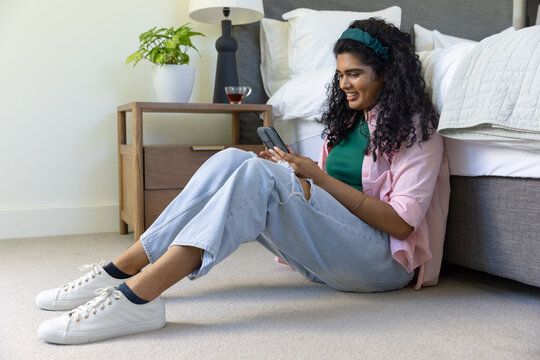 Indian woman sitting on bedroom floor, wearing pink shirt and ripped jeans, holding smartphone