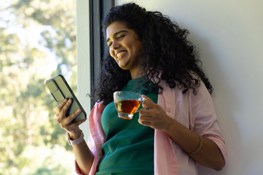 Indian woman leaning by window, smiling while holding phone and tea, in pink shirt and bangles