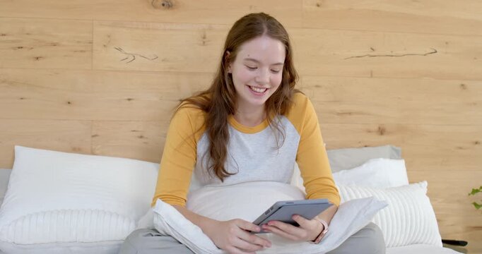 Adult female smiling while looking at tablet, sitting on bed, tapping and reading, wearing raglan