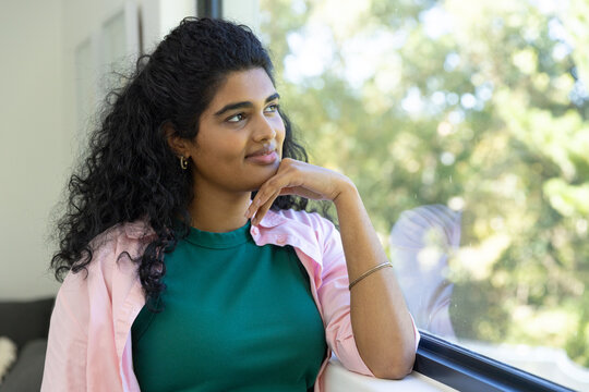 Indian woman sitting at home by large window, resting chin, wearing green top and pink overshirt