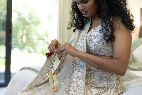 Indian female sitting on bed adjusting beaded tassel while wearing ornate dupatta near glass door