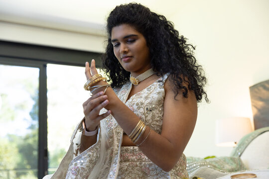 Indian woman sitting on bed by sliding glass door, adjusting gold bangles with choker and saree